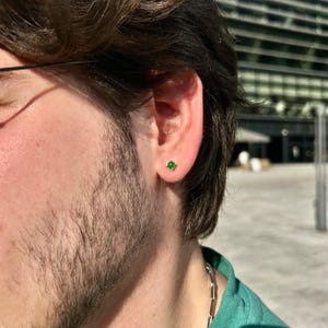 Puede incluir: Primer plano de una oreja con un peque&ntilde;o pendiente de bot&oacute;n cuadrado verde esmeralda. El pendiente est&aacute; engastado en un marco dorado o plateado. La persona lleva una camisa verde y una cadena de plata.