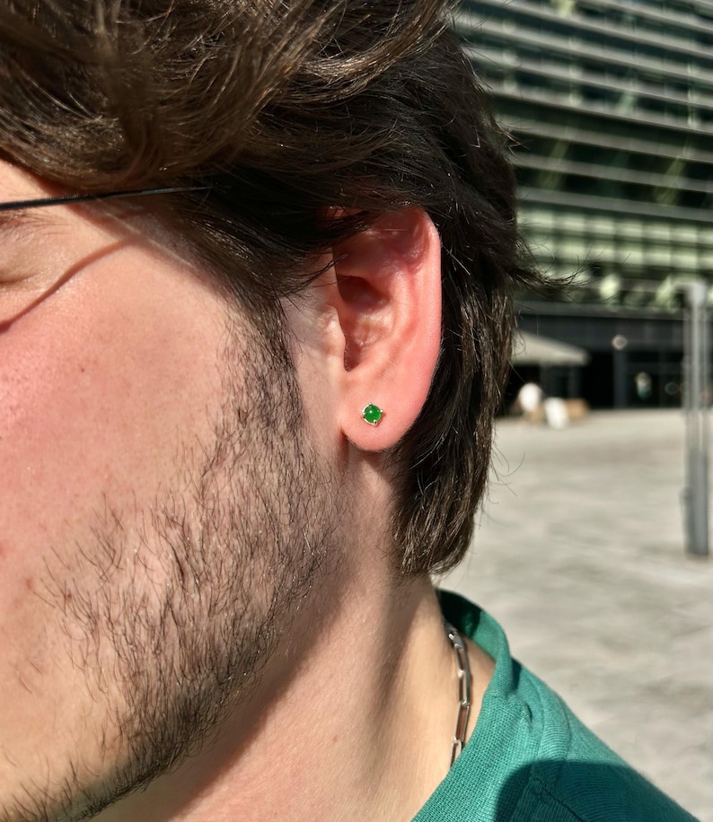 Puede incluir: Primer plano de una oreja con un peque&ntilde;o pendiente de bot&oacute;n cuadrado verde esmeralda. El pendiente est&aacute; engastado en un marco dorado o plateado. La persona lleva una camisa verde y una cadena de plata.