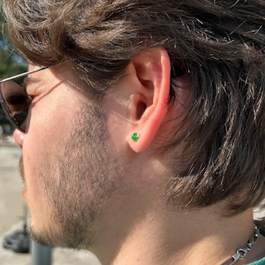 Puede incluir: Primer plano de la oreja de una persona con un peque&ntilde;o pendiente de bot&oacute;n de piedra preciosa verde de forma cuadrada. El pendiente est&aacute; engastado en metal dorado o plateado. La persona lleva gafas de sol y tiene el pelo oscuro y barba.