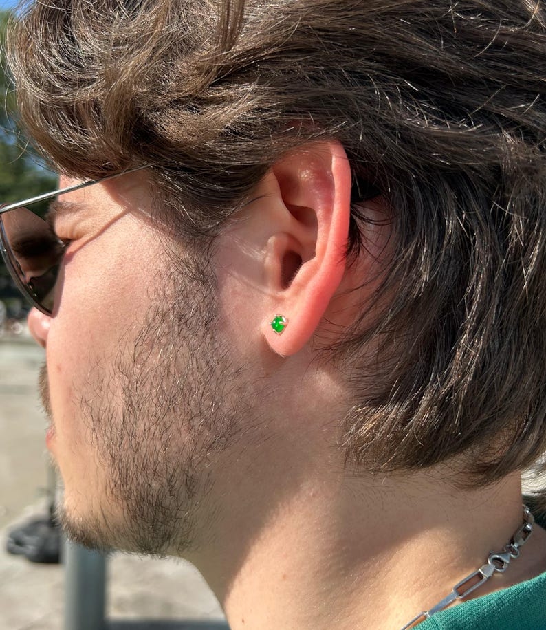 Puede incluir: Primer plano de la oreja de una persona con un peque&ntilde;o pendiente de bot&oacute;n de piedra preciosa verde de forma cuadrada. El pendiente est&aacute; engastado en metal dorado o plateado. La persona lleva gafas de sol y tiene el pelo oscuro y barba.