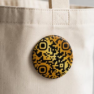 May include: A round pinback button featuring a black and gold QR code design attached to a beige canvas tote bag. The button has a metallic gold gradient background with black square pixel patterns forming a scannable code structure.