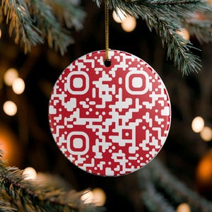 May include: A round, flat ceramic ornament featuring a red and white QR code pattern. The ornament has a small hole at the top for hanging and is suspended by a thin gold-colored string. The design consists of a dense, pixelated grid of red and white.