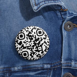 May include: A circular pin-back button featuring a black and white QR code pattern. The button is attached to a blue denim fabric surface with visible stitching and a dark metallic button detail, creating a high-contrast graphic accessory.