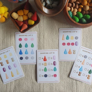 May include: Five colorful cards with shopping lists for a game. The lists include numbers, squares, and circles with different colored shapes inside. The cards are on a beige surface.