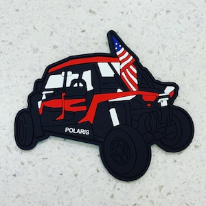May include: Black and red Polaris off-road vehicle with a white American flag attached to the roll cage.