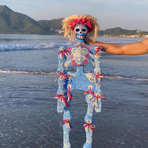 May include: A blue articulated skeleton figure featuring a painted skull with a floral crown and blonde hair. The body is decorated with white scrollwork and red and white ribbon bows at each joint, creating a detailed and colorful appearance.