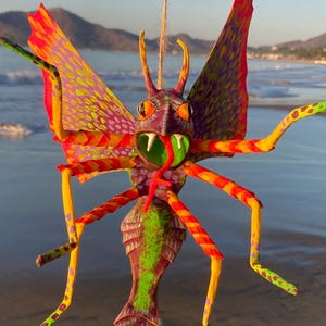 May include: A vibrant, handcrafted insect sculpture with a colorful design. The artwork features a detailed head, wings, and legs in shades of red, orange, yellow, green, and purple. It is suspended by a natural fiber string, perfect for hanging.