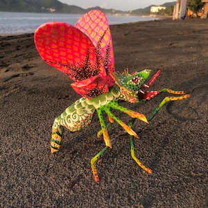 May include: A vibrant, hand-painted wooden insect sculpture with intricate patterns. The wings are predominantly red and pink, and the body and legs are decorated with green, yellow, and orange. The artwork is displayed on a dark sandy beach with the sea in the background.