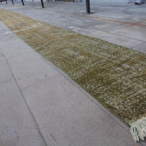 Puede incluir: Una alfombra de pasillo larga, verde y blanca con flecos en los extremos. La alfombra está colocada sobre un suelo de hormigón.