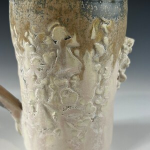 May include: A ceramic mug with a light brown glaze and a white glaze with a floral design. The mug has a handle and a textured surface.