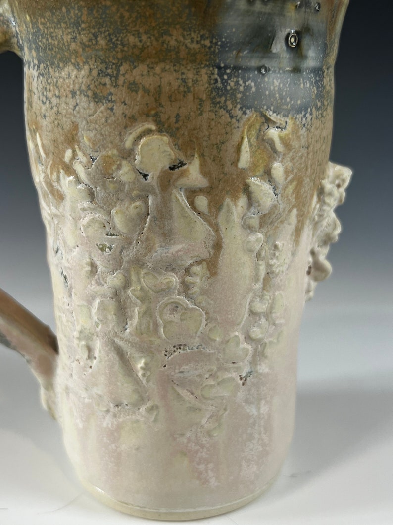 May include: A ceramic mug with a light brown glaze and a white glaze with a floral design. The mug has a handle and a textured surface.