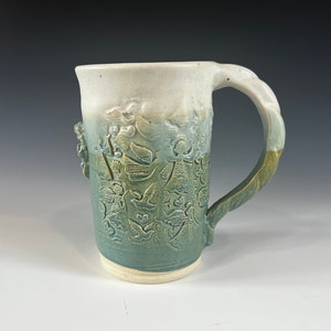 May include: A ceramic mug with a green and white glaze. The mug has an embossed design of flowers and leaves. The handle is thick and rounded.