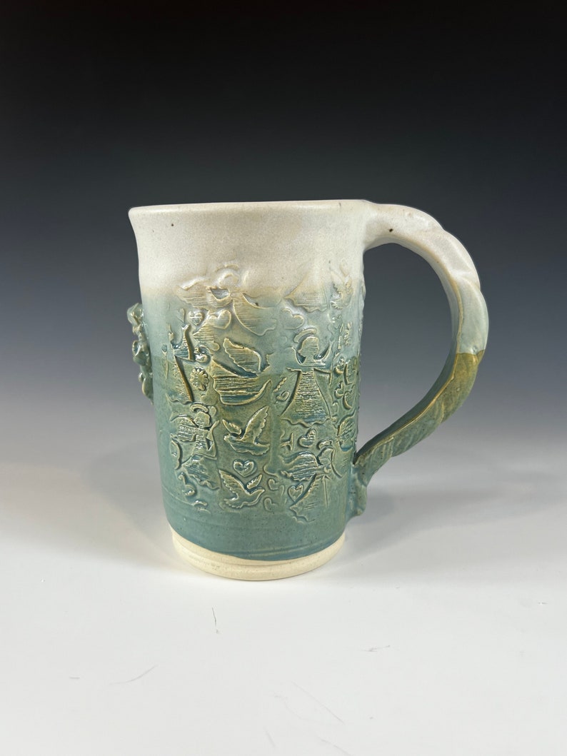 May include: A ceramic mug with a green and white glaze. The mug has an embossed design of flowers and leaves. The handle is thick and rounded.