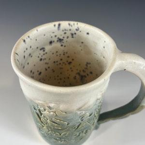 May include: A ceramic mug with a green and white glaze. The mug has a speckled white interior and a handle. The exterior of the mug has a raised design of leaves and flowers.