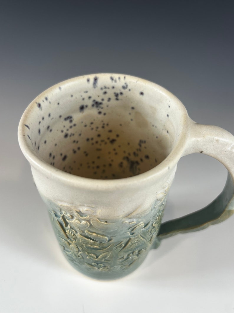 May include: A ceramic mug with a green and white glaze. The mug has a speckled white interior and a handle. The exterior of the mug has a raised design of leaves and flowers.