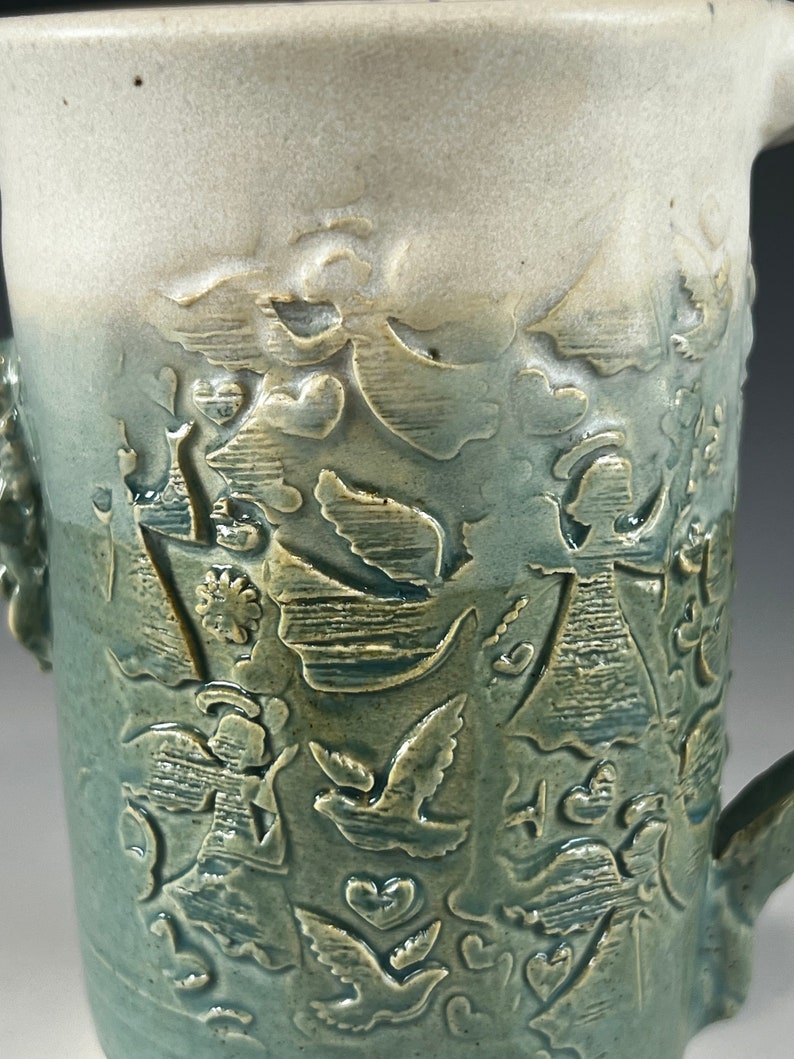 May include: A ceramic mug with a green and white glaze. The mug has a raised design of angels and doves.
