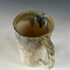 May include: A ceramic mug with a textured, speckled glaze in shades of brown, white, and gray. The mug has a large handle and a wide, rounded opening.