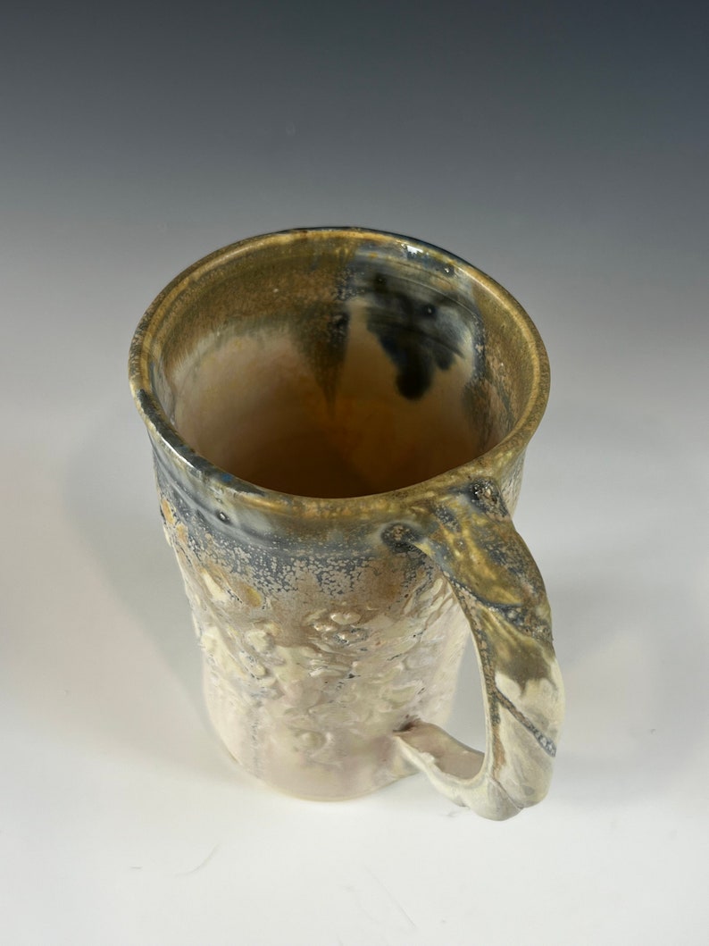 May include: A ceramic mug with a textured, speckled glaze in shades of brown, white, and gray. The mug has a large handle and a wide, rounded opening.