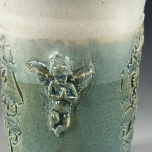 May include: A ceramic mug with a light blue glaze and a white glaze on the top. The mug has a raised design of an angel with wings and a praying pose. The angel is in a light yellow color.