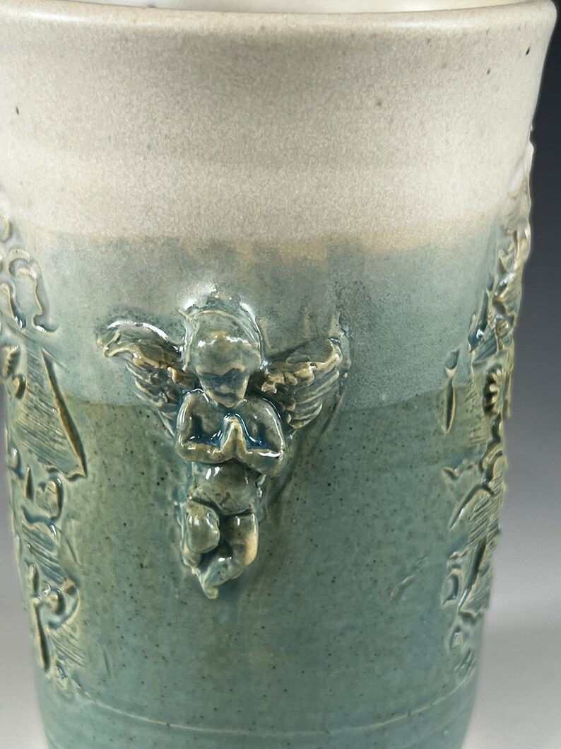 May include: A ceramic mug with a light blue glaze and a white glaze on the top. The mug has a raised design of an angel with wings and a praying pose. The angel is in a light yellow color.