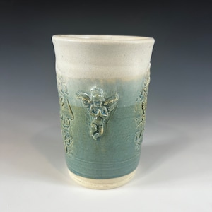 May include: A white ceramic mug with a blue glaze and a raised design of angels. The mug has a white rim and base.