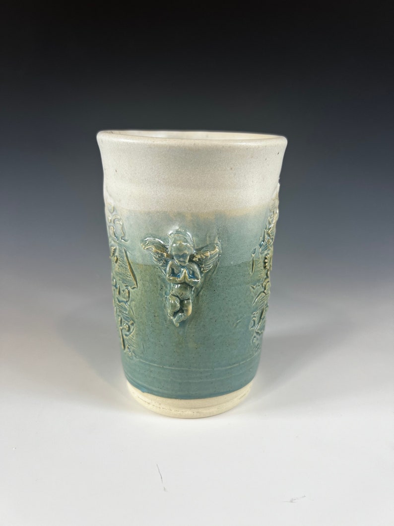 May include: A white ceramic mug with a blue glaze and a raised design of angels. The mug has a white rim and base.