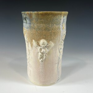 May include: A ceramic vase with a light brown, white, and blue glaze. The vase has a textured surface with raised designs of angels.