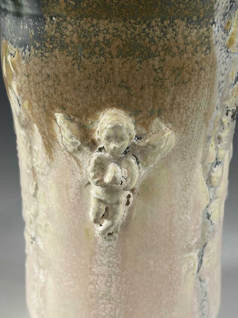 May include: A ceramic vase with a textured, speckled glaze in shades of brown, gray, and white. The vase features a raised, sculpted angel design.