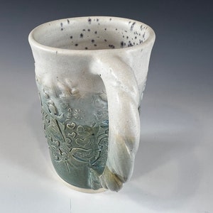 May include: A white ceramic mug with a green and blue glaze. The mug has a unique, textured design with a raised floral pattern. The handle is thick and twisted.