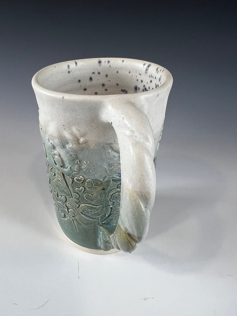 May include: A white ceramic mug with a green and blue glaze. The mug has a unique, textured design with a raised floral pattern. The handle is thick and twisted.