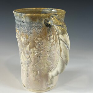 May include: A ceramic mug with a light brown glaze and a white floral design. The mug has a unique, sculpted handle.