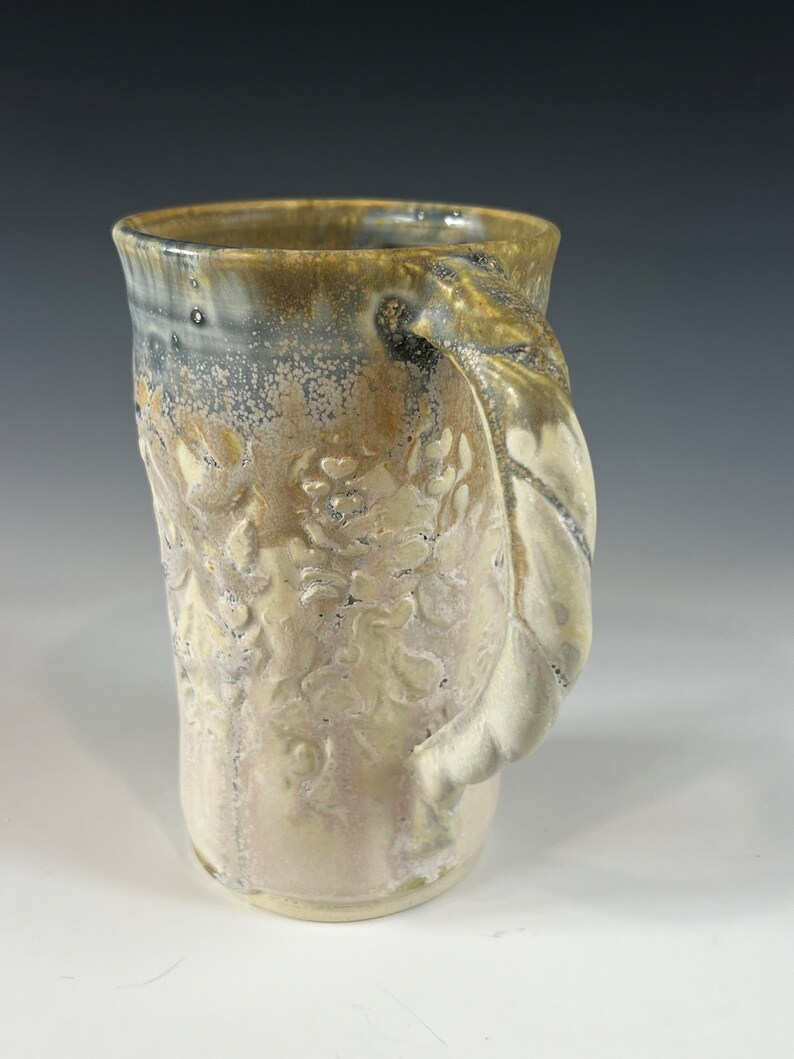 May include: A ceramic mug with a light brown glaze and a white floral design. The mug has a unique, sculpted handle.