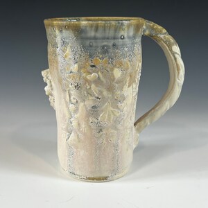 May include: A white ceramic mug with a textured, floral design and a brown handle. The mug has a speckled glaze and a slightly uneven surface.