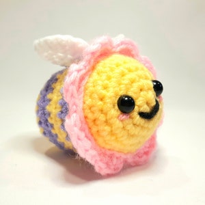 Peut inclure: Un jouet d'abeille en crochet avec un corps jaune, un abdomen rayé de violet et de jaune, des ailes en forme de fleur rose et des yeux noirs. L'abeille a un visage souriant.