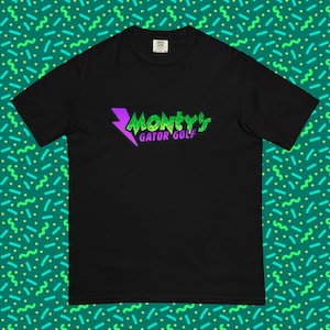 Puede incluir: Camiseta negra con el texto "Monty's Gator Golf" en verde y morado. El diseño incluye un rayo morado y una fuente verde y morada. La camiseta es de manga corta y cuello redondo.