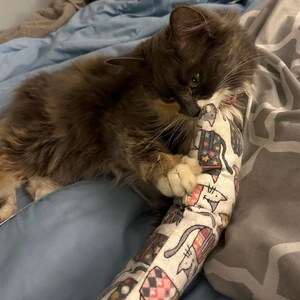 May include: A long, cylindrical cat toy made of white fabric featuring a repeating pattern of illustrated cats wearing colorful hats and scarves. The toy is being held by a fluffy, long-haired gray and white cat with dark facial markings.