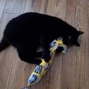 May include: A solid black cat playing with a long, yellow, cylindrical fabric toy featuring a repeating pattern of cartoon animal faces, including owls and other creatures, with a small grey fabric tag attached to the bottom end.
