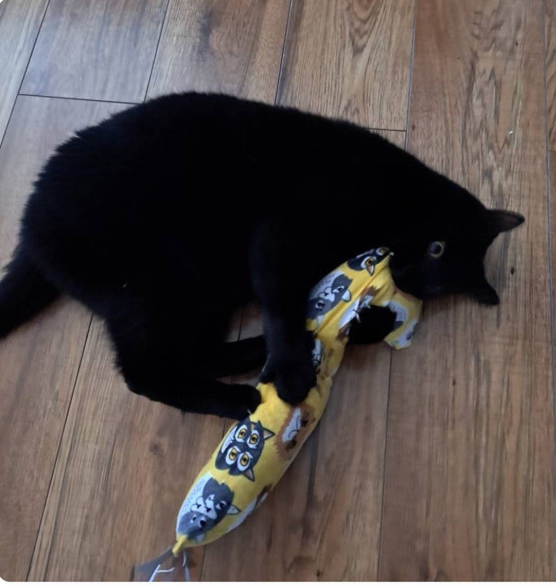May include: A solid black cat playing with a long, yellow, cylindrical fabric toy featuring a repeating pattern of cartoon animal faces, including owls and other creatures, with a small grey fabric tag attached to the bottom end.