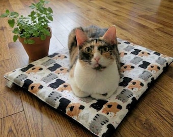 May include: A calico cat sits on a rectangular pet mat featuring a repeating pattern of small black, orange, and grey cat illustrations on a white background. The mat has a white fabric edge and a small white tag on the side.