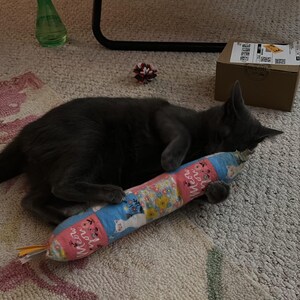 May include: A solid gray cat lying down and holding a cylindrical fabric toy. The toy features a repeating pattern of pink panels with the text 'meow you' and blue panels decorated with illustrations of white cats and colorful floral arrangements.