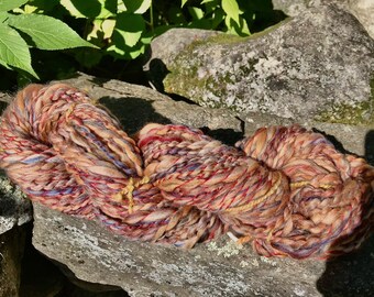 May include: A skein of yarn in a mix of brown, red, blue, and gold colors. The yarn is thick and fluffy, and it is arranged in a loose coil.