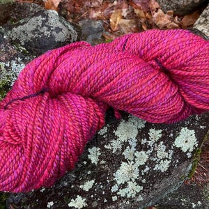 May include: A skein of pink and red yarn with a variegated color pattern. The yarn is twisted and piled on a rock surface.