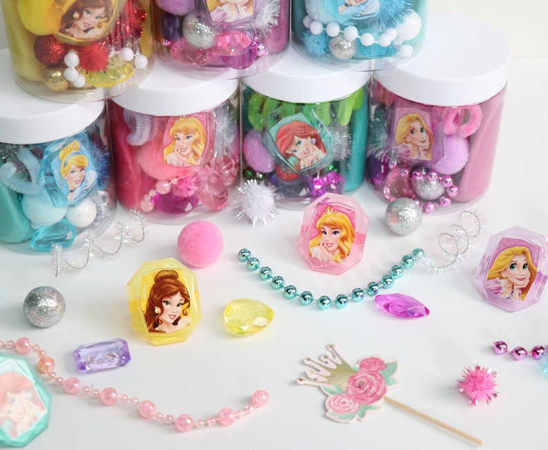 May include: A collection of princess-themed party favor jars filled with colorful beads, pom poms, and other small trinkets. The jars are arranged in a pyramid shape, with the top jar being a light blue color and the bottom jar being a yellow color. There are also several princess-themed rings and necklaces scattered around the jars.