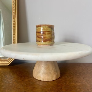 May include: A small, cylindrical ceramic cup with horizontal brown and yellow bands sits on a white marble cake stand with a light wood base. The stand is on a dark wood surface. A gold framed mirror is in the background.