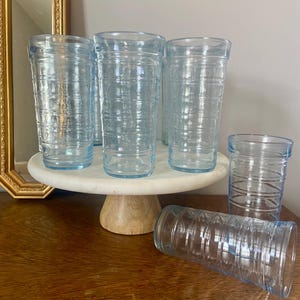 May include: A collection of clear, light blue drinking glasses with a horizontal ribbed design. The glasses are arranged on a white marble cake stand with a wooden base. One glass is lying on a wooden surface.