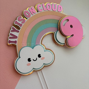 May include: A layered paper cake topper featuring a pastel rainbow with a smiling white cloud at the base and a large pink number nine on the right side. The text IVY IS ON CLOUD is arched in pink letters over the rainbow, all outlined in gold foil.