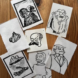 May include: A collection of black and white linocut prints on textured paper. The prints depict various subjects, including a garlic bulb, a skeleton, and cartoon characters. The prints are arranged on a wooden surface.