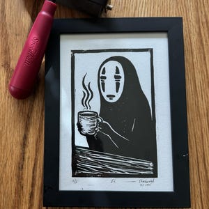 Puede incluir: Impresión artística enmarcada en negro de un personaje sosteniendo una taza con vapor ascendente. La impresión está firmada y fechada. Una rodillo Speedball rojo y una herramienta para tallar madera también están en la imagen.