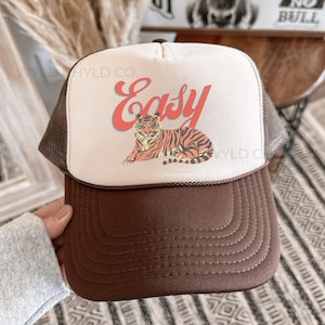 May include: A brown and white trucker hat with a brown mesh back. The front of the hat features a white tiger with red text that says "Easy".