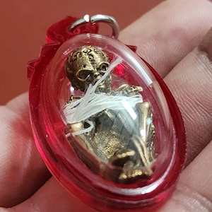May include: A small, gold-colored figurine of a skeleton wrapped in white string, encased in a clear, oval-shaped plastic pendant with a red border. The pendant has a silver-colored metal loop at the top.